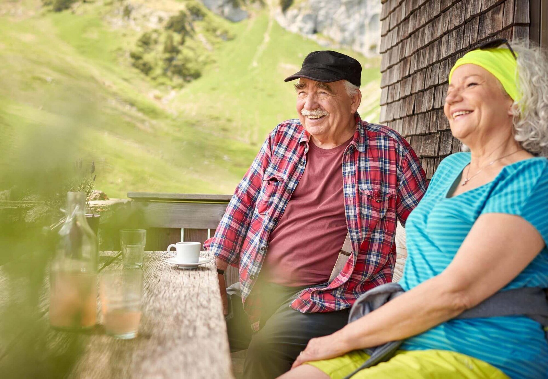 Ein Ehepaar sitzt an einem Tisch an einer Berghütte und macht eine Pause. Sie schauen beide freudig in die Ferne. Ausreichend Bewegung regt die Durchblutung an und hilft dabei, der Entstehung einer Erkrankung von Diabetes mellitus Typ 2 entgegenzuwirken.