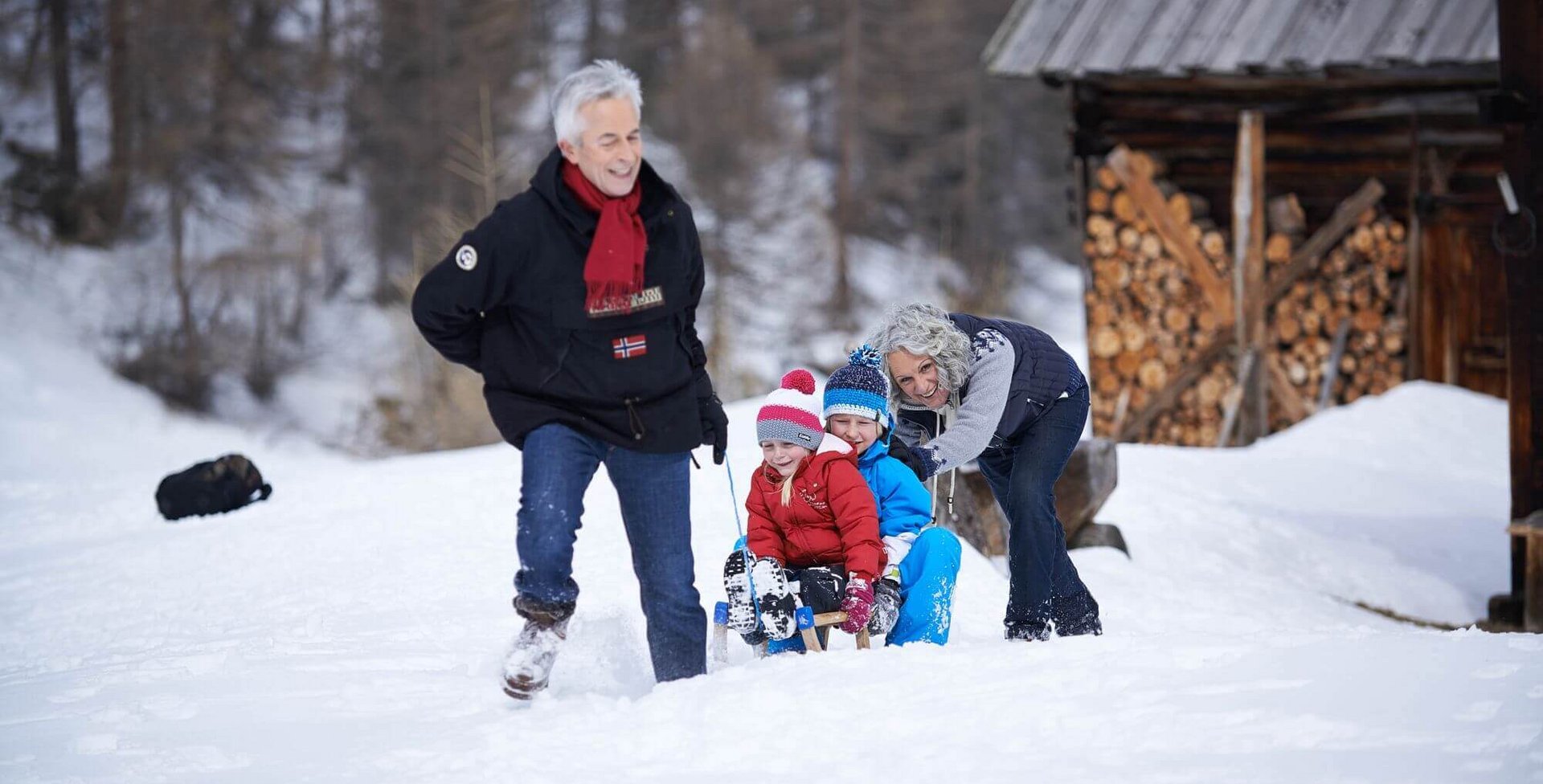 Ein älteres Pärchen zieht zwei Kinder, welche auf einem Schlitten sitzen, durch den Schnee. Schon einfache Bewegungen wie Bücken, Drehen oder Aufrichten können das Lendenwirbelsäulen-Syndrom (kurz: LWS-Syndrom) auslösen.