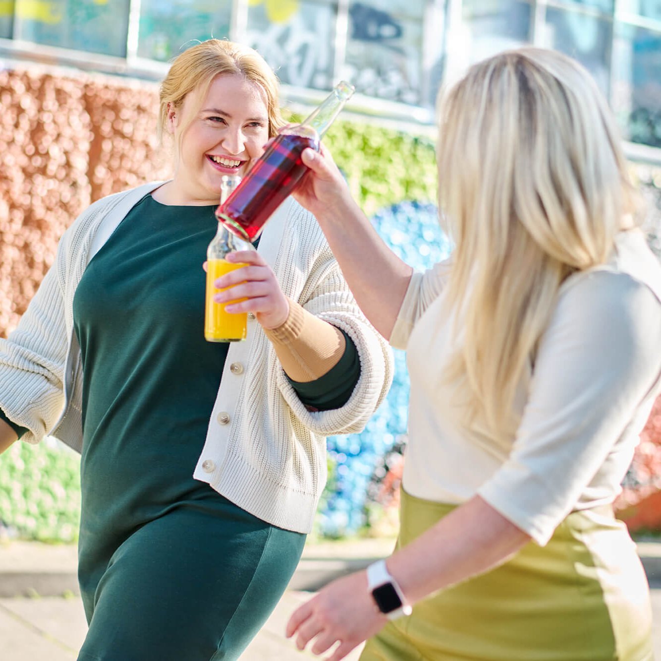 Zwei Frauen stoßen mit farbenfrohen Getränkeflaschen an, während sie lächelnd durch eine urbane Umgebung spazieren. Im Hintergrund ist eine bunte Wand mit grafischen Mustern zu sehen, die eine lebendige Atmosphäre schafft.
