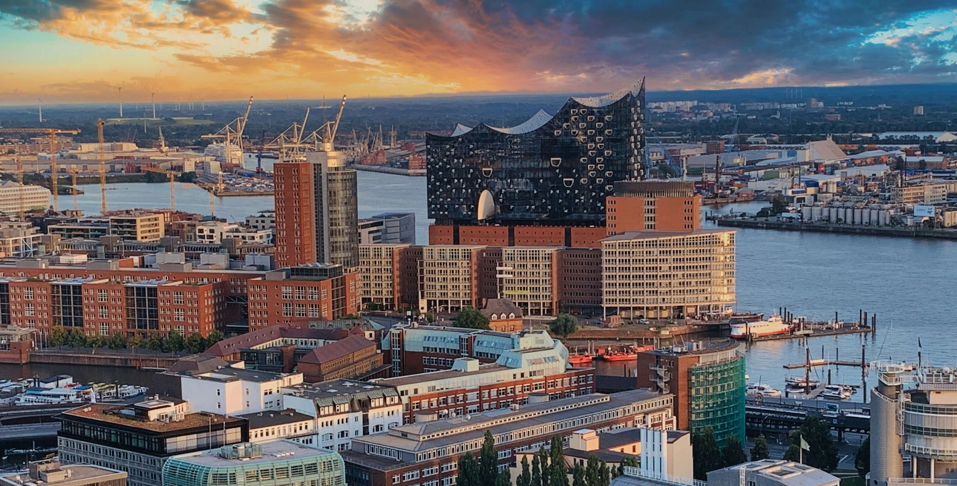 Panoramablick auf Hamburg mit der Elbphilharmonie im Zentrum, umgeben von modernen und historischen Gebäuden entlang der Elbe, im Vordergrund eine dichte Bebauung bei Sonnenuntergang mit dramatischem Himmel.