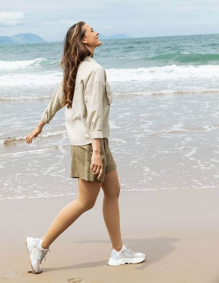 Eine Frau trägt VenoTrain soft Kompressionsstrümpfe und spaziert entspannt am Strand entlang. Sie trägt ein beiges Hemd, eine kurze olivgrüne Hose und weiße Sneakers. 