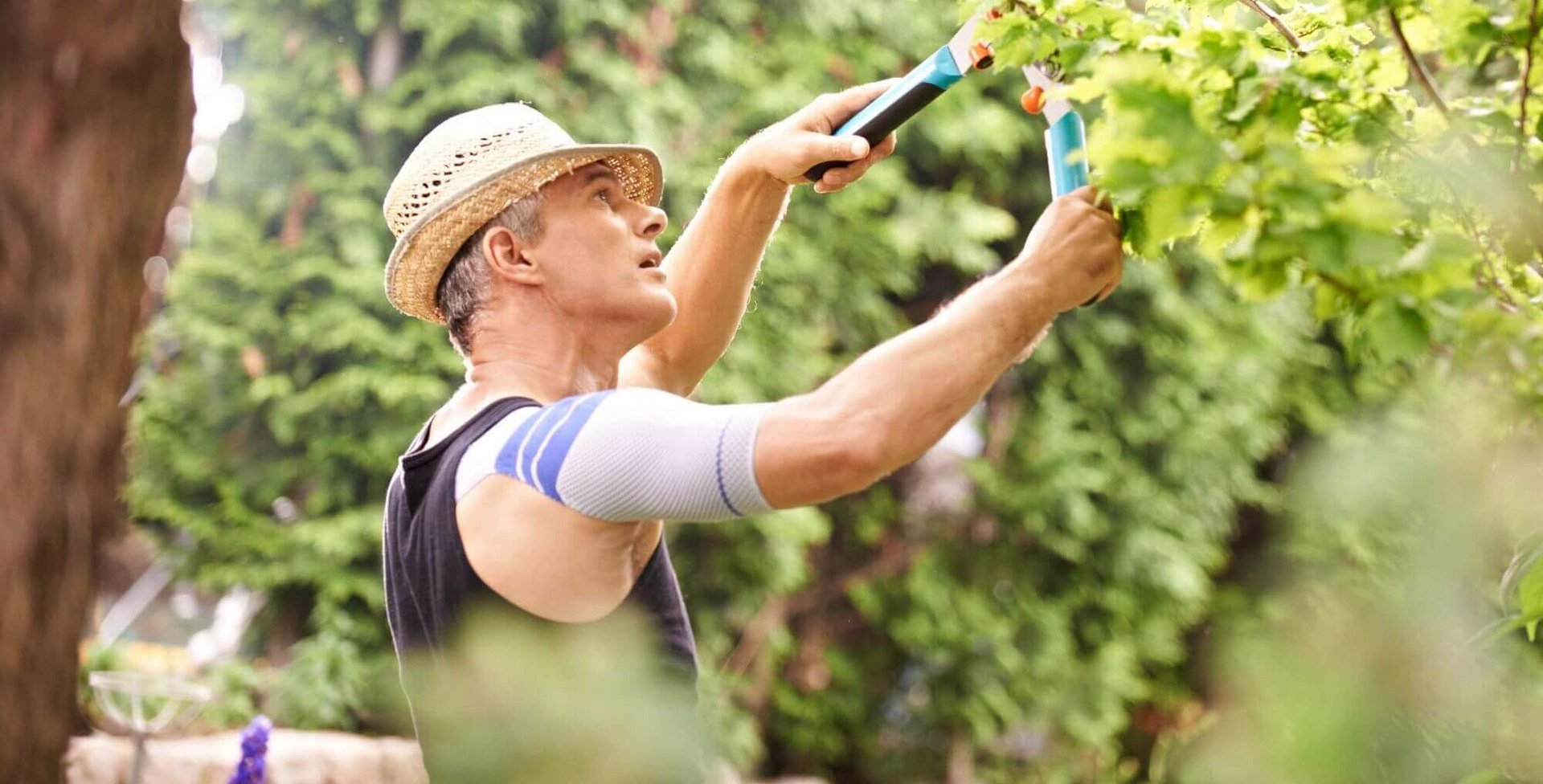 Auf dem Bild ist ein Mann zu sehen, der mit einer Heckenschere im Garten arbeitet. Er trägt die OmaTrain Schulterbandage von Bauerfeind, die eine stabilisierende Wirkung hat und die Führung des Gelenks verbessert.