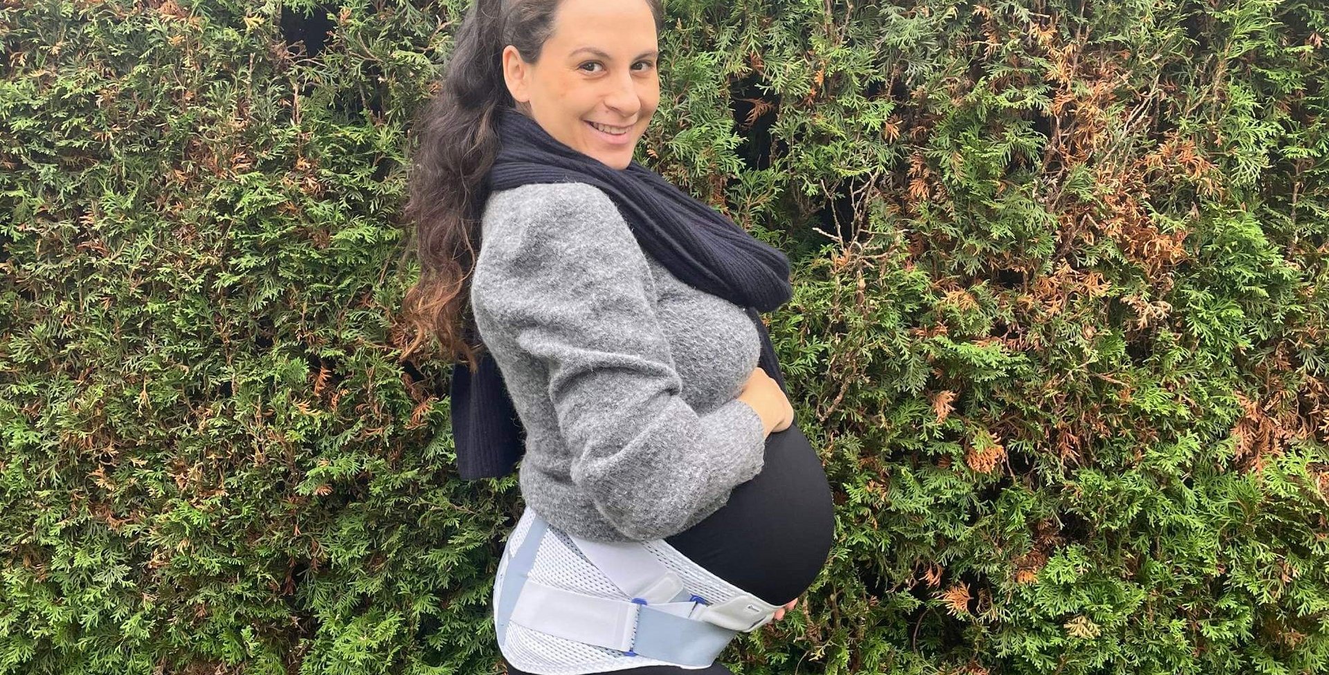 Schwangere Frau steht im Profil vor einer Hecke, trägt eine graue Jacke, schwarzen Schal und eine weiße Schwangerschaftsorthese um den Bauch.