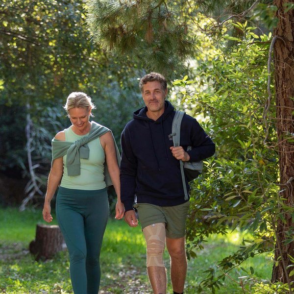 Das Bild zeigt einen jungen Mann und seine Partnerin, die zusammen durch einen Wald wandern. Der Mann trägt dabei die GenuTrain Bandage von Bauerfeind zur Stabilisierung seines Knies.