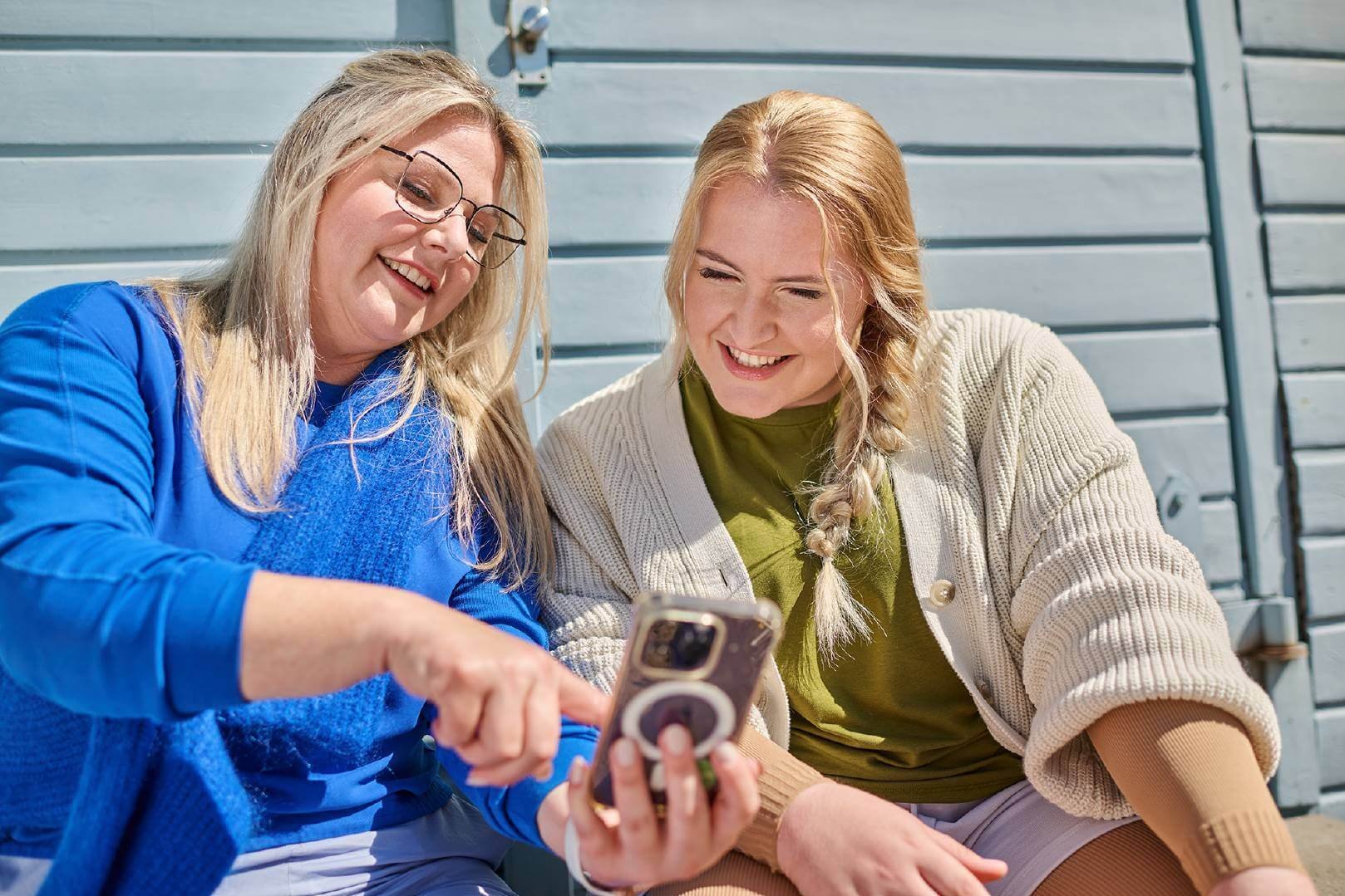 Zwei Frauen sitzen draußen vor einer blauen Holzwand und lächeln, während sie gemeinsam auf ein Smartphone schauen. 