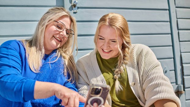 Zwei Frauen sitzen draußen vor einer blauen Holzwand und lächeln, während sie gemeinsam auf ein Smartphone schauen. 