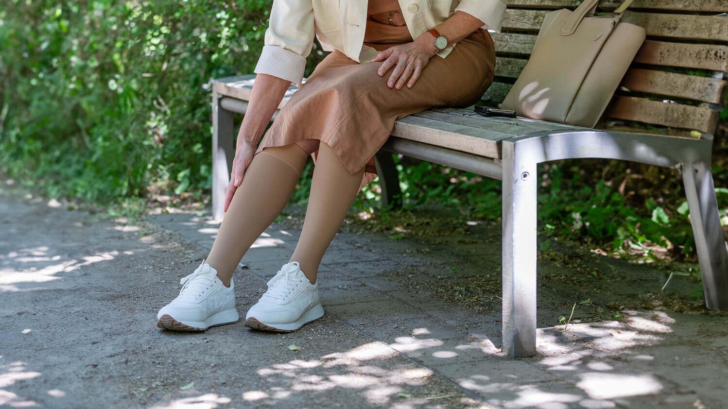 Bildausschnitt einer Frau auf einer Bank. Sie fasst sich mit ihrer rechten Hand an ihre rechte Wade. Vorübergehende Schwellungen der Füße, Knöchel oder Unterschenkel können erste Warnzeichen einer chronisches Venenschwäche (chronische venöse Insuffizienz, CVI) sein.