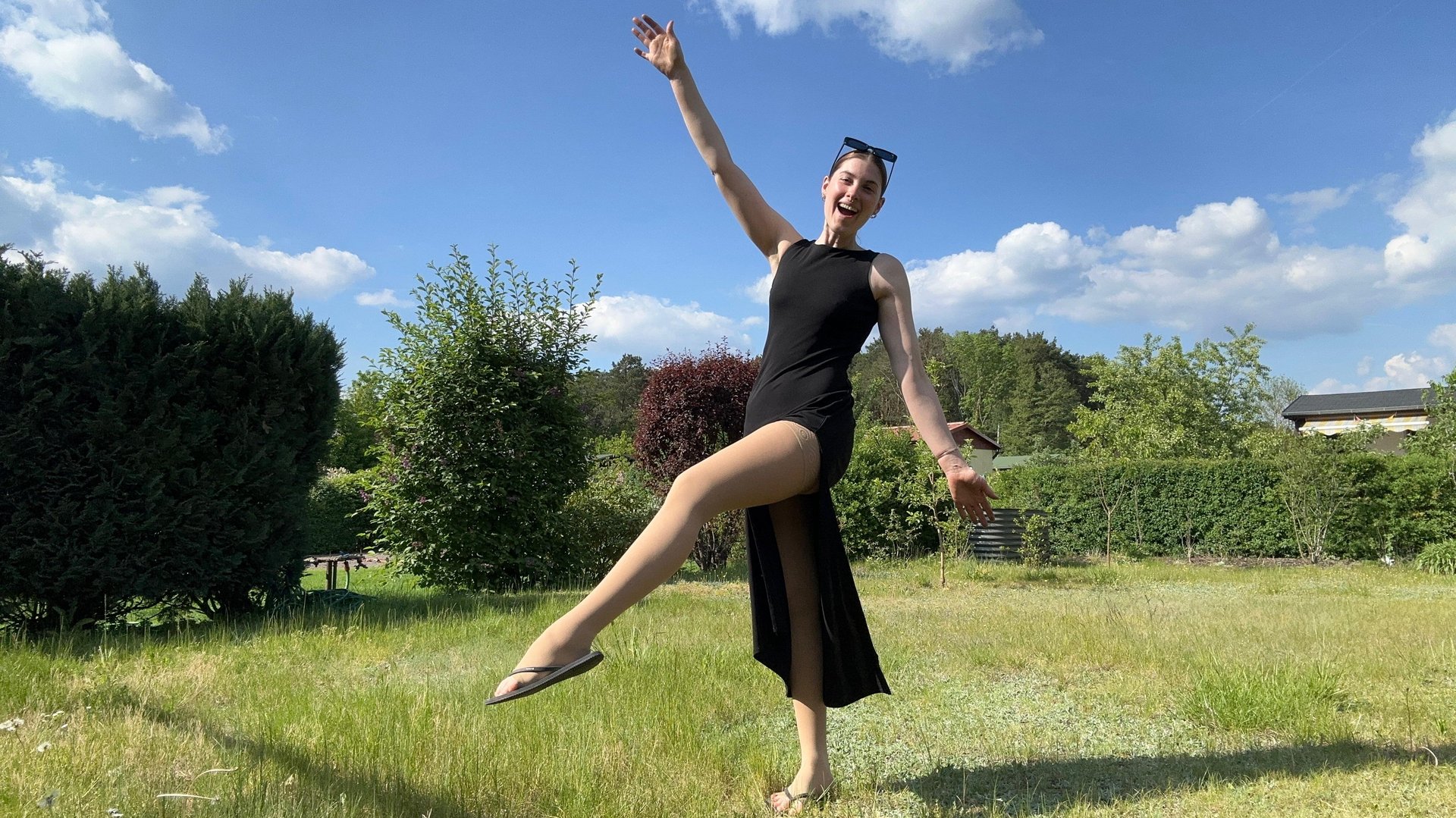 Eva Seemann steht in einem Garten und hebt lachend ein Bein. Sie trägt ein schwarzes Kleid und Bauerfeind Kompressionsstrümpfe.