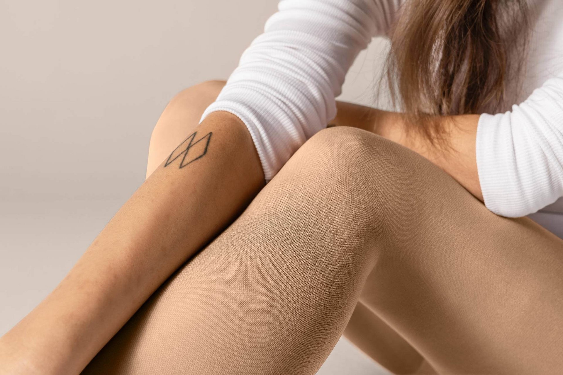 Detailaufnahme von beigen Strumpfhosen und verschränkten Armen einer sitzenden Frau mit weissem Oberteil und geometrischem Tattoo am Unterarm. Detailaufnahme von beigen Strumpfhosen und verschränkten Armen einer sitzenden Frau mit weissem Oberteil und geometrischem Tattoo am Unterarm.