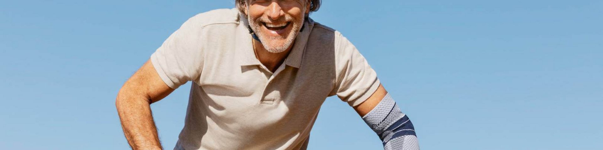 Mann mit Fahrradhelm fährt bei klarem Himmel lächelnd Fahrrad, trägt die EpiTrain Ellenbogenbandage von Bauerfeind am linken Arm zur Unterstützung des Gelenks