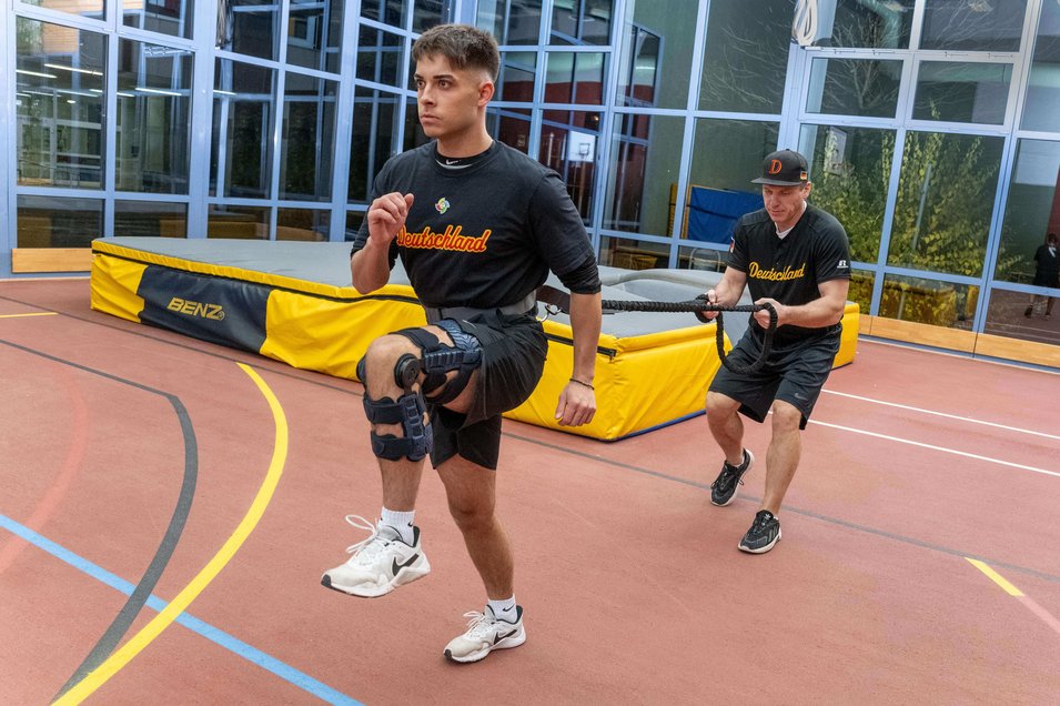 Simon Liedkte, Baseballspieler, führt ein Widerstandstraining in einer Halle durch. Er trägt die SecuTec Genu Flex Knieorthese am rechten Bein, um die Stabilität nach seinem hinteren Kreuzbandriss zu gewährleisten. Hinter ihm hält ein Trainer ein Widerstandsseil, das an Simons Hüfte befestigt ist.