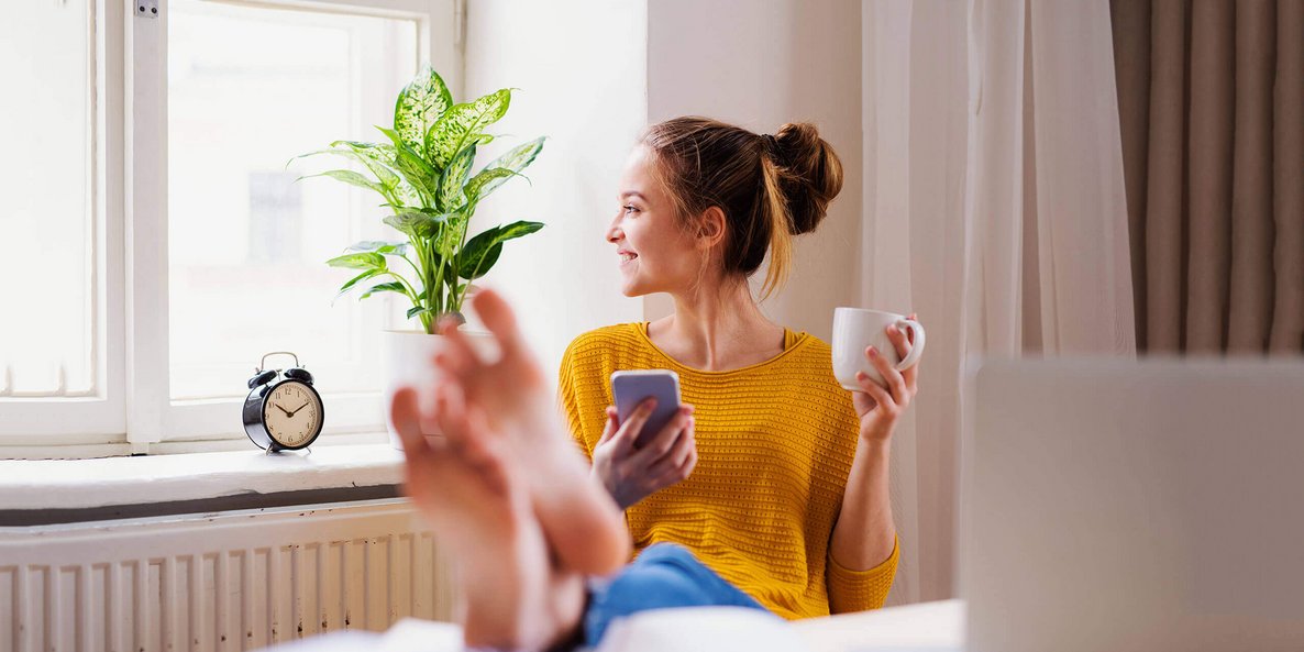 Auf dem Bild sitzt eine junge Frau auf einem Stuhl und hat ihre Füße auf den Tisch vor ihr abgelegt. In der linken Hand hält sie eine Tasse; in der rechten Hand ein Smartphone. Regelmäßiges Training der Fußmuskulatur kann Schmerzen vorbeugen.