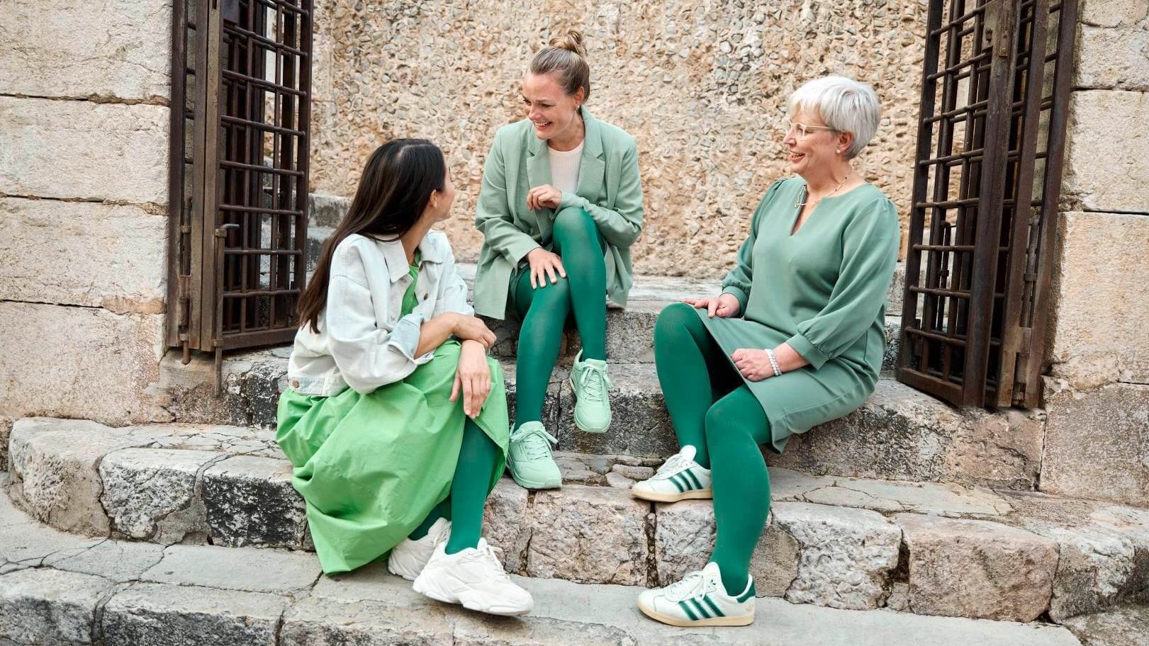 Drei Frauen sitzen auf historischen Steinstufen vor einer rustikalen Steinmauer und unterhalten sich lachend. Sie tragen Outfits in verschiedenen Grüntönen, ergänzt durch VenoTrain Kompressionsstrümpfe in der Modefarbe 'Irish Green'.
