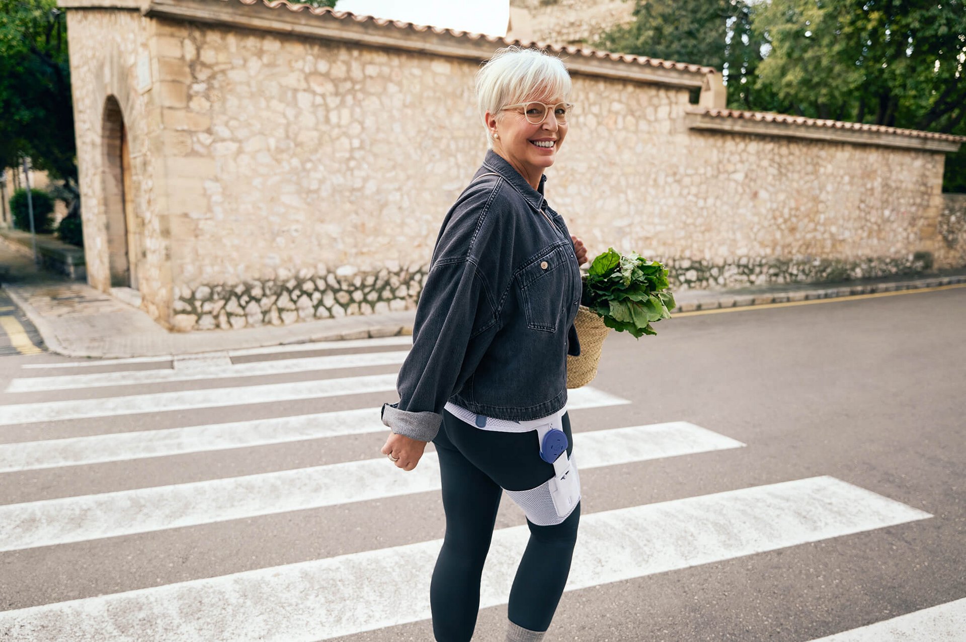 Eine ältere Frau mit kurzen weissen Haaren und Brille überquert einen Zebrastreifen. Sie trägt eine Jeansjacke, dunkle Leggings mit medizinischen Geräten daran und hält einen Korb mit frischem Grünzeug. Im Hintergrund ist eine Steinmauer mit Dachziegeln sowie Bäume zu sehen.