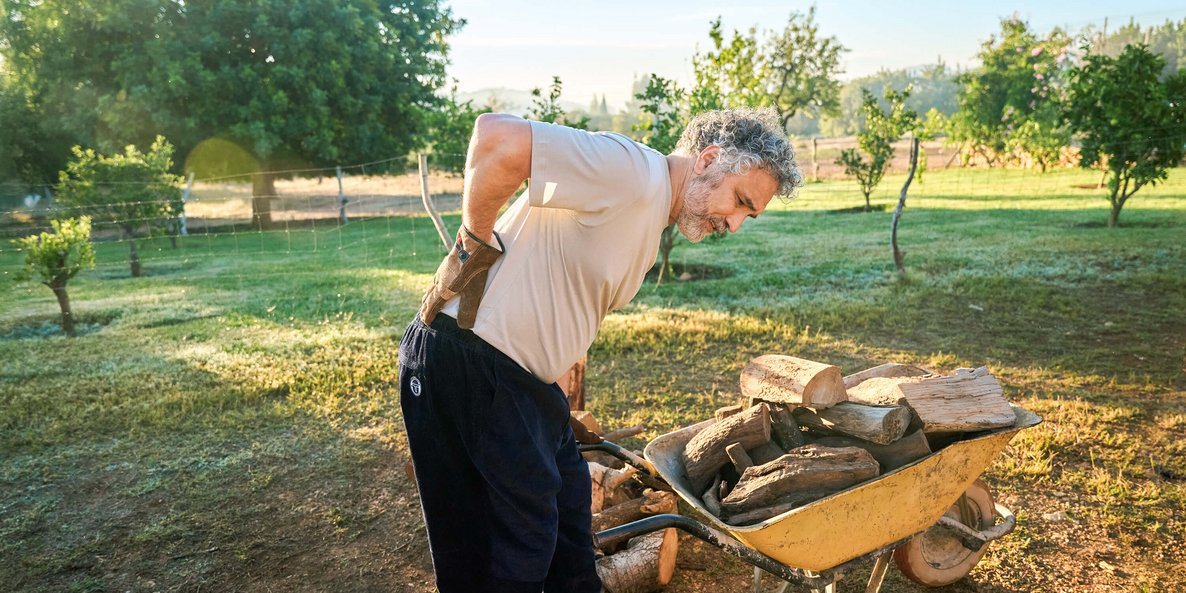 Ein älterer Mann mit grauen Haaren hält sich im Garten den schmerzenden Rücken, während er sich über eine Schubkarre mit Holzscheiten beugt. Im Hintergrund sind grüne Bäume und ein ländlicher Garten zu sehen.