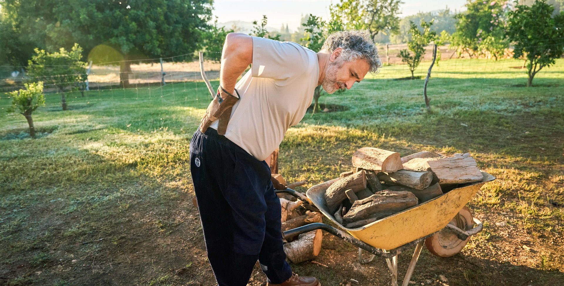 Ein älterer Mann mit grauen Haaren hält sich im Garten den schmerzenden Rücken, während er sich über eine Schubkarre mit Holzscheiten beugt. Im Hintergrund sind grüne Bäume und ein ländlicher Garten zu sehen.