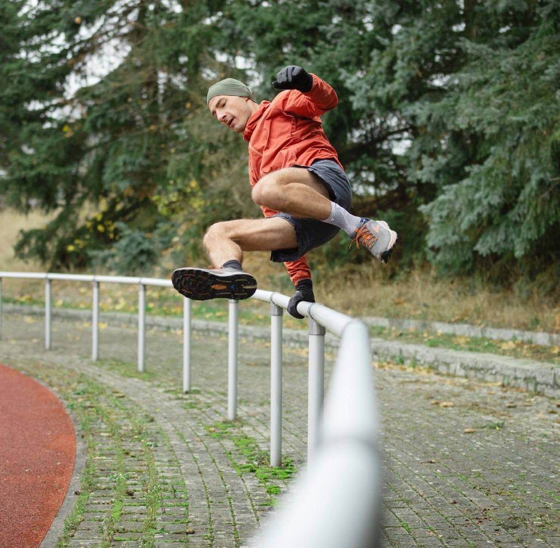 Ein sportlicher Mann springt mit Schwung über eine Geländerstange auf einer Laufbahn. Er trägt die AchilloTrain Bandage von Bauerfeind, die seine Achillessehne unterstützt.