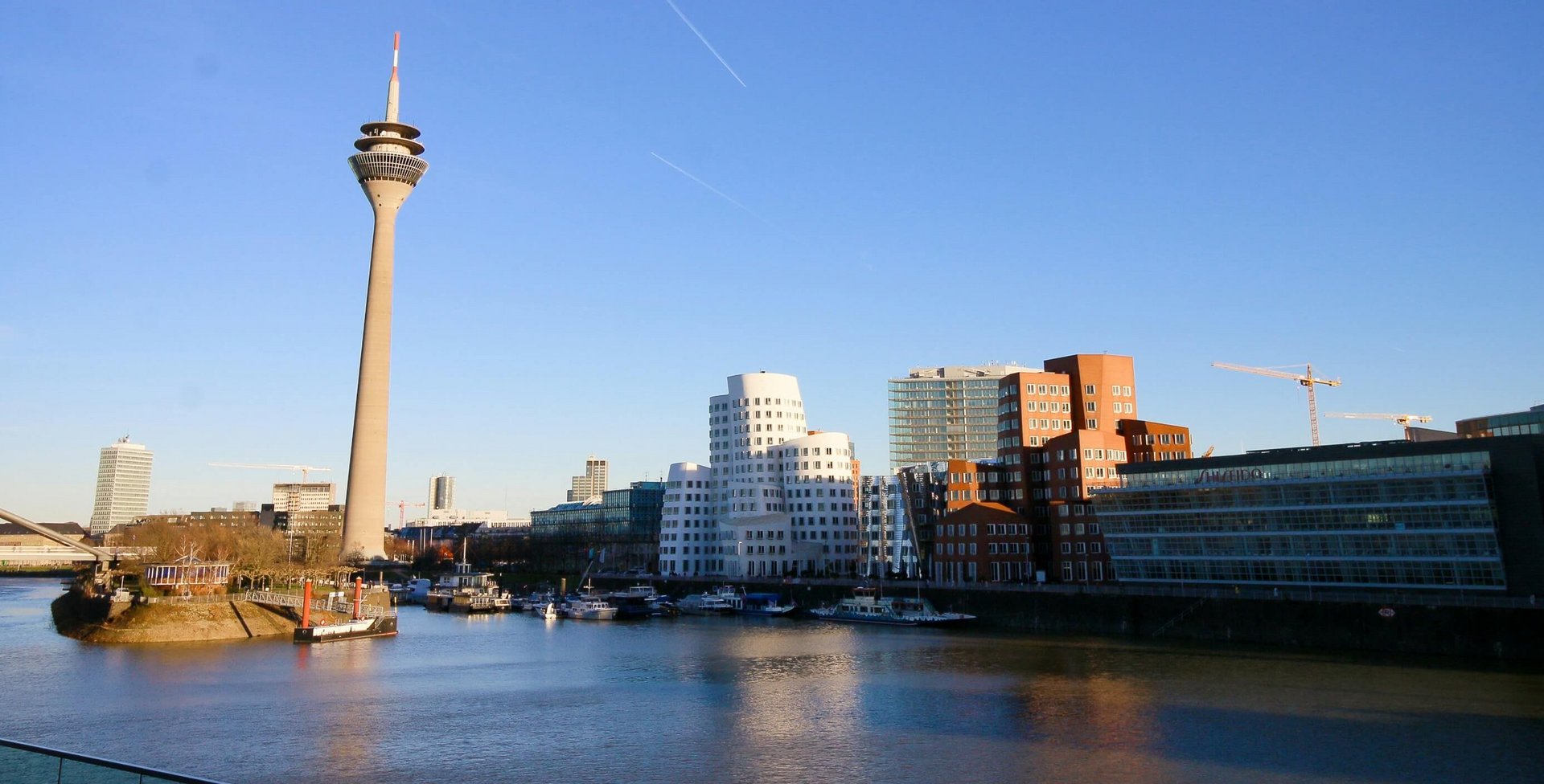 Blick auf den MedienHafen in Düsseldorf mit dem Rheinturm links, modernen Bürogebäuden entlang des Hafenbeckens und klar blauem Himmel, einige Boote im Wasser und Baukräne im Hintergrund.