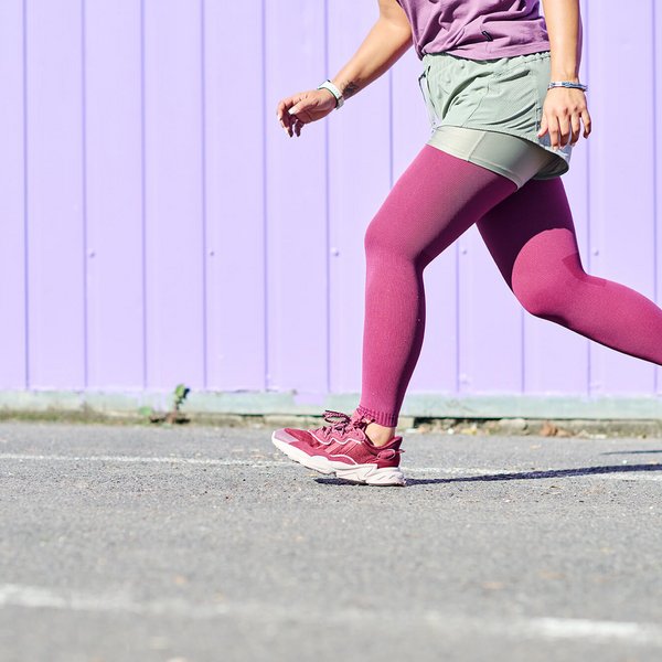 Frau in sportlicher Kleidung mit bordeauxfarbenen Strumpfhosen und passenden Turnschuhen läuft auf Asphalt vor einer lilafarbenen Holzwand. Nur der Unterkörper und ein Teil des Oberkörpers sind sichtbar.