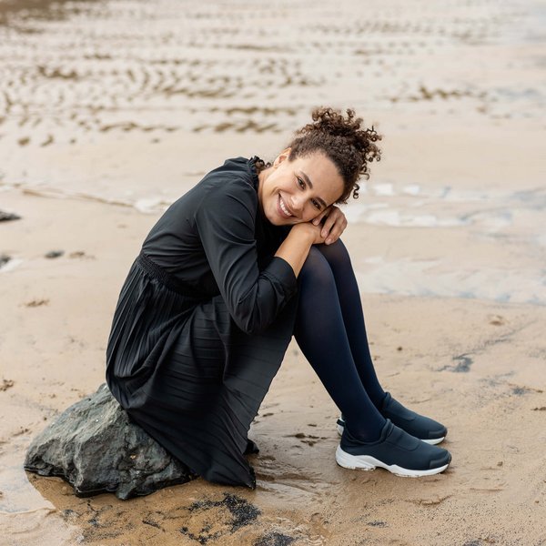 Das Bild zeigt eine Frau die auf einem kleinen Felsen am Strand sitzt. Sie trägt VenoTrain soft Kompressionsstrümpfe von Bauerfeind.