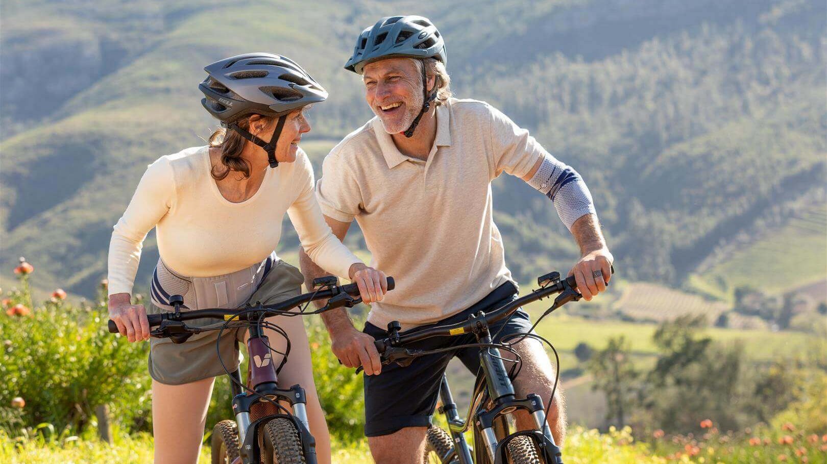 Abgebildet ist ein älteres Paar, die auf einer Radtour unterwegs sind. Um Kreuzschmerzen vorzubeugen, trägt die Frau eine LumboTrain Rückenbandage von Bauerfeind.
