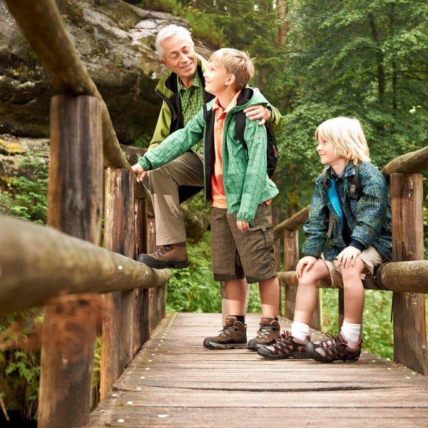 Ein älterer Herr läuft mit zwei Kindern über eine Holzbrücke. Das Tragen von Kindereinlagen dient zur Behandlung von Schmerzen, Fehlstellungen und neurologischen Erkrankungen.