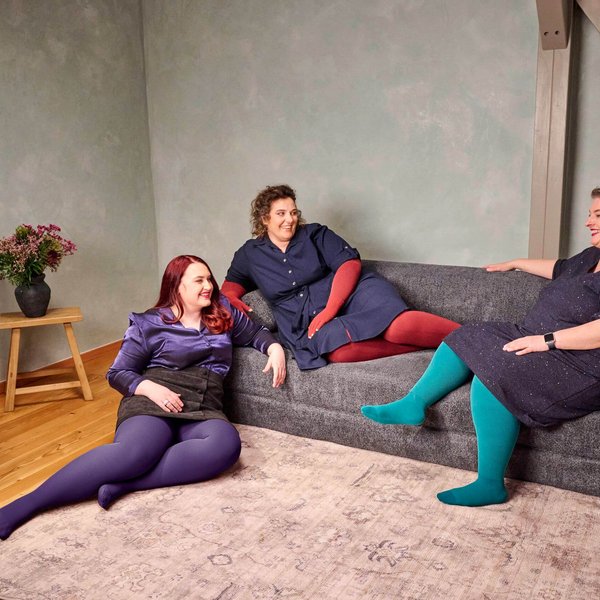 Drei Frauen sitzen und liegen entspannt auf einem dunkelgrauen Sofa in einem modern eingerichteten Wohnzimmer mit Holzfussboden. Sie tragen farbenfrohe Strumpfhosen – in Violett, Rot und Türkis – kombiniert mit dunklen Kleidern oder Röcken. Alle wirken fröhlich und unterhalten sich miteinander. Links steht ein kleiner Holztisch mit einer schwarzen Vase und einem Blumenstrauss.