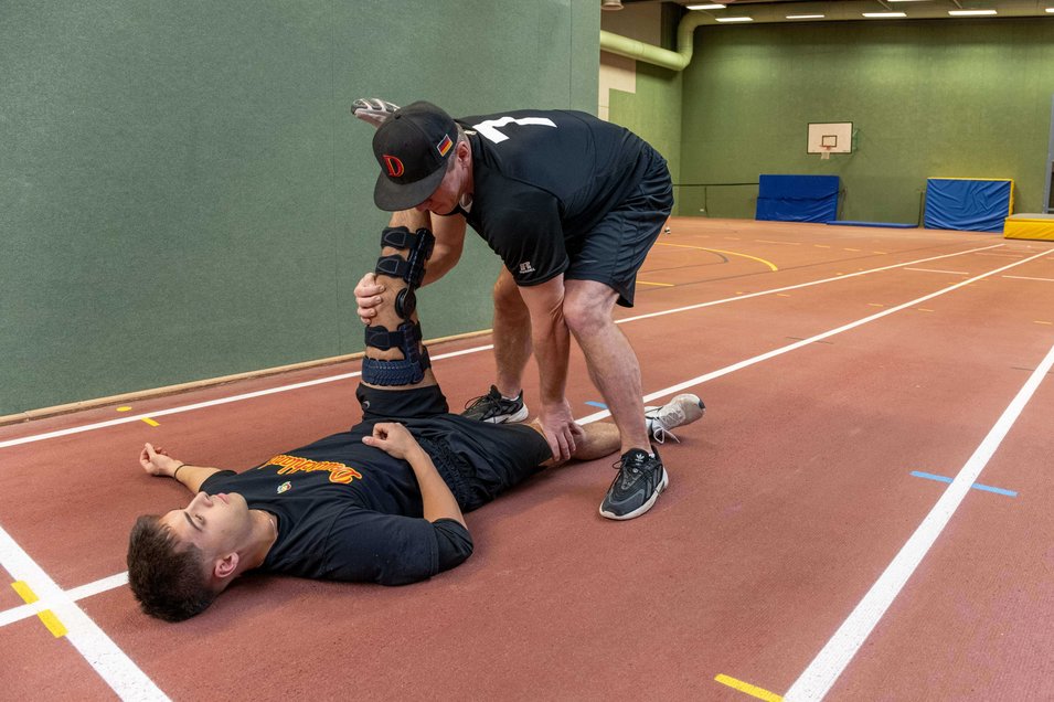 Baseballspieler Simon Liedkte liegt während einer Reha-Übung in einer Sporthalle auf dem Boden. Sein Bein wird von einem Trainer gedehnt, während er eine SecuTec Genu Flex Knieorthese trägt, um das Gelenk nach einem hinteren Kreuzbandriss zu stabilisieren.