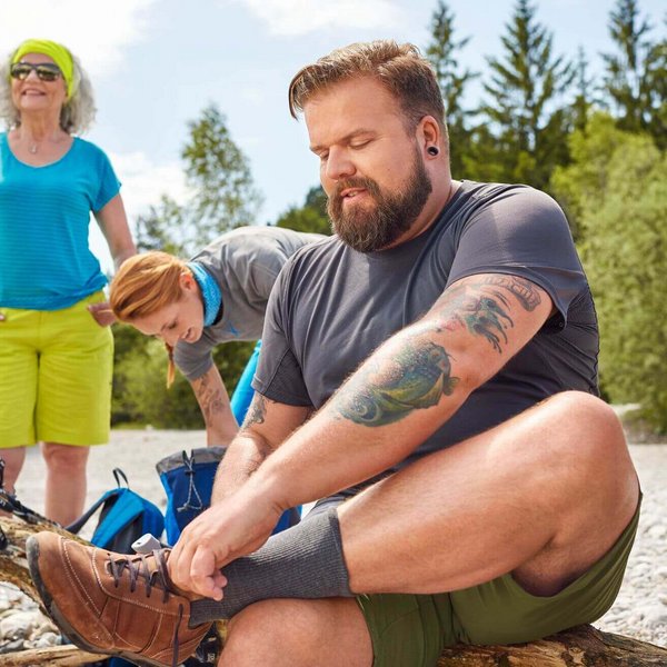 Eine Wandergruppe macht einen Rast auf einem Baumstamm. Der Mann in der Mitte des Bildes schnürt seinen linken Wanderschuh zu. Hochwertige Weichpolster-Einlagen wie die ErgoPad soft Diabetes von Bauerfeind helfen Diabetikern dabei, Druckstellen und Wunden an den Füßen während des Laufens zu vermeiden.