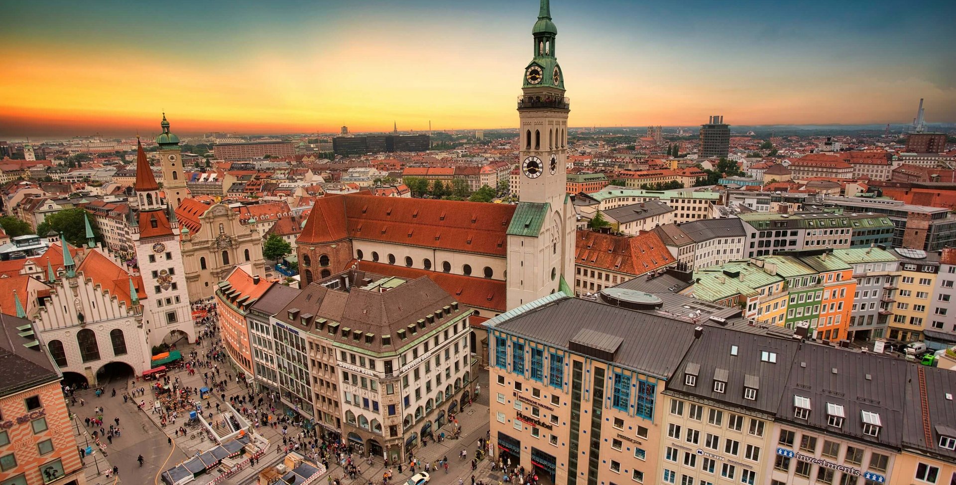 Luftaufnahme vom Marienplatz in München mit der Peterskirche im Zentrum, umgeben von historischen Gebäuden mit roten Dächern und zahlreichen Menschen auf dem Platz bei Sonnenuntergang.