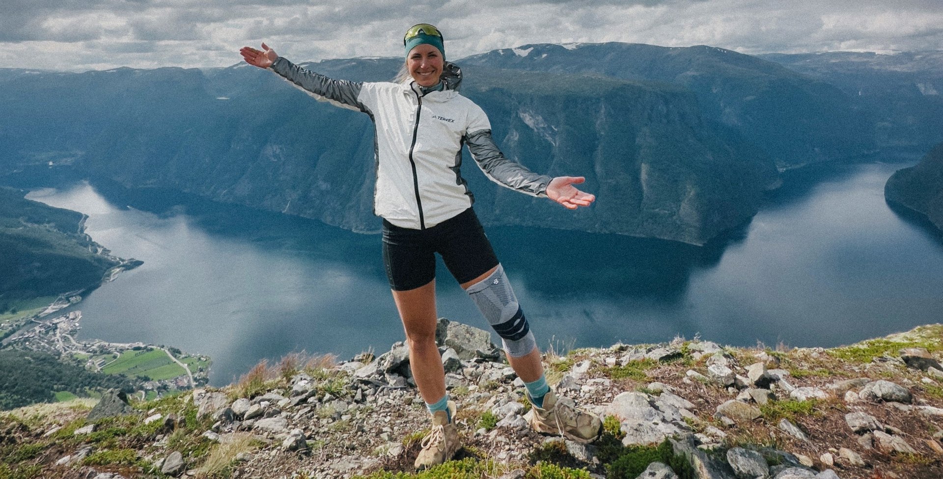Antonia Horn steht lächelnd auf einem Berggipfel mit ausgebreiteten Armen. Sie trägt eine GenuTrain Kniebandage von Bauerfeind am rechten Knie. Im Hintergrund erstreckt sich eine atemberaubende Landschaft mit Fjorden und Bergen unter einem teils bewölkten Himmel.