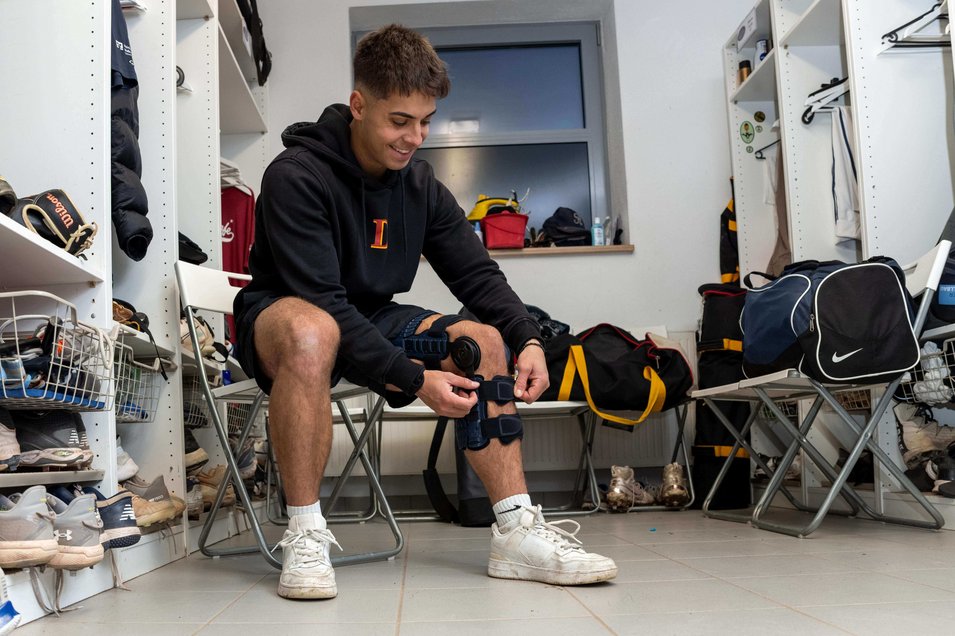 Simon Liedkte sitzt in der Umkleidekabine und legt sich die SecuTec Genu Flex Knieorthese an. Um ihn herum sind Sporttaschen, Schuhe und Baseballausrüstung zu sehen. Er lächelt konzentriert, während er die Orthese anpasst.
