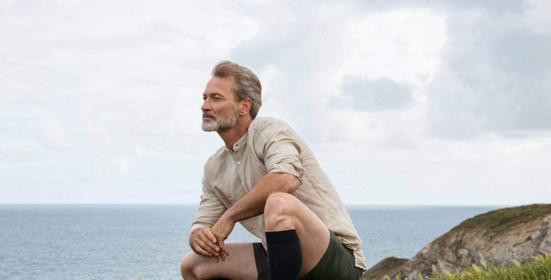Ein Mann hockt auf einer Wiese am Meer. Er trägt Kompressionsstrümpfe von Bauerfeind.