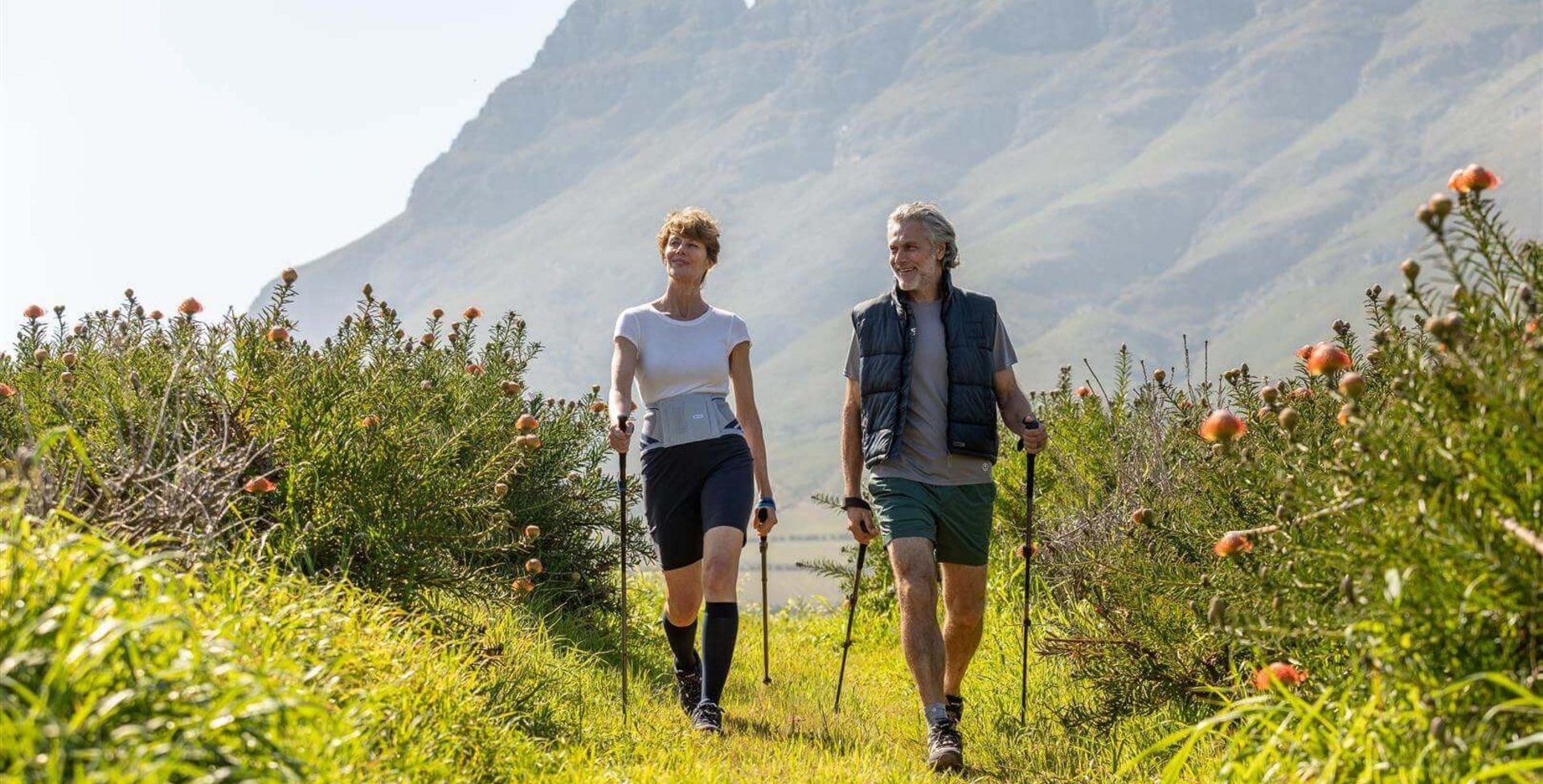 Auf dem Bild kann man sehen, wie ein älteres Paar in der Natur Nordic Walking betreibt. Sie tragen Train-Bandagen von Bauerfeind, um ihre Gelenke und den Rücken während der Bewegung zu entlasten.