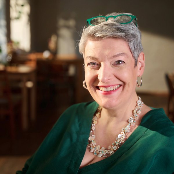 Porträt einer lächelnden Frau mit kurzem grauen Haar, grüner Brille auf dem Kopf, grüner Bluse und auffälliger Halskette, sitzend in einem gemütlichen Café mit Holzmöbeln im Hintergrund.