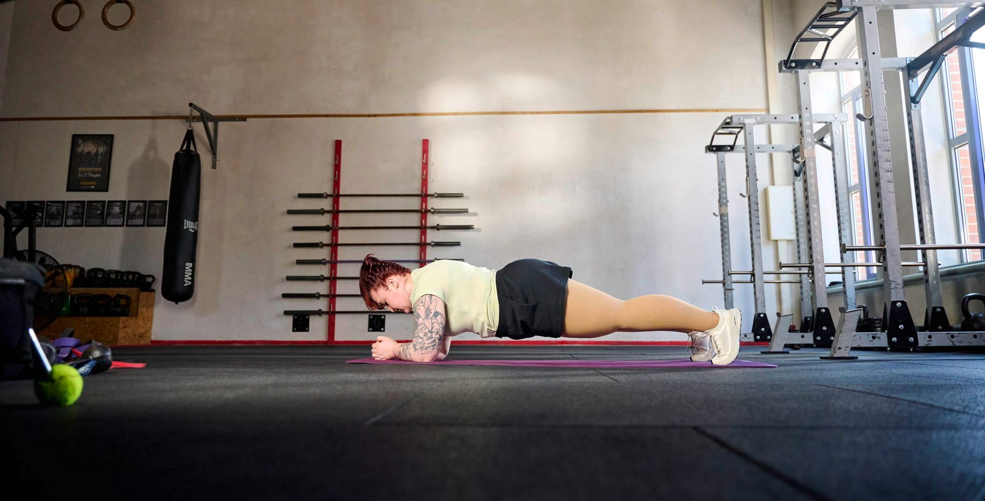 Person macht eine Unterarmstütz-Übung (Plank) auf einer Fitnessmatte in einem geräumigen Trainingsraum mit hohen Decken, Hantelstangen an der Wand und Fenstern auf der rechten Seite.