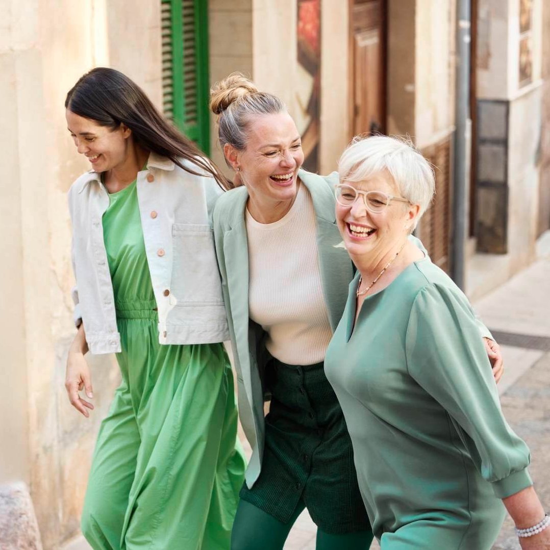 Drei lachende Frauen in stilvollen grünen Outfits spazieren Arm in Arm durch eine sonnige Altstadtstraße, symbolisieren Lebensfreude und Gemeinschaft.