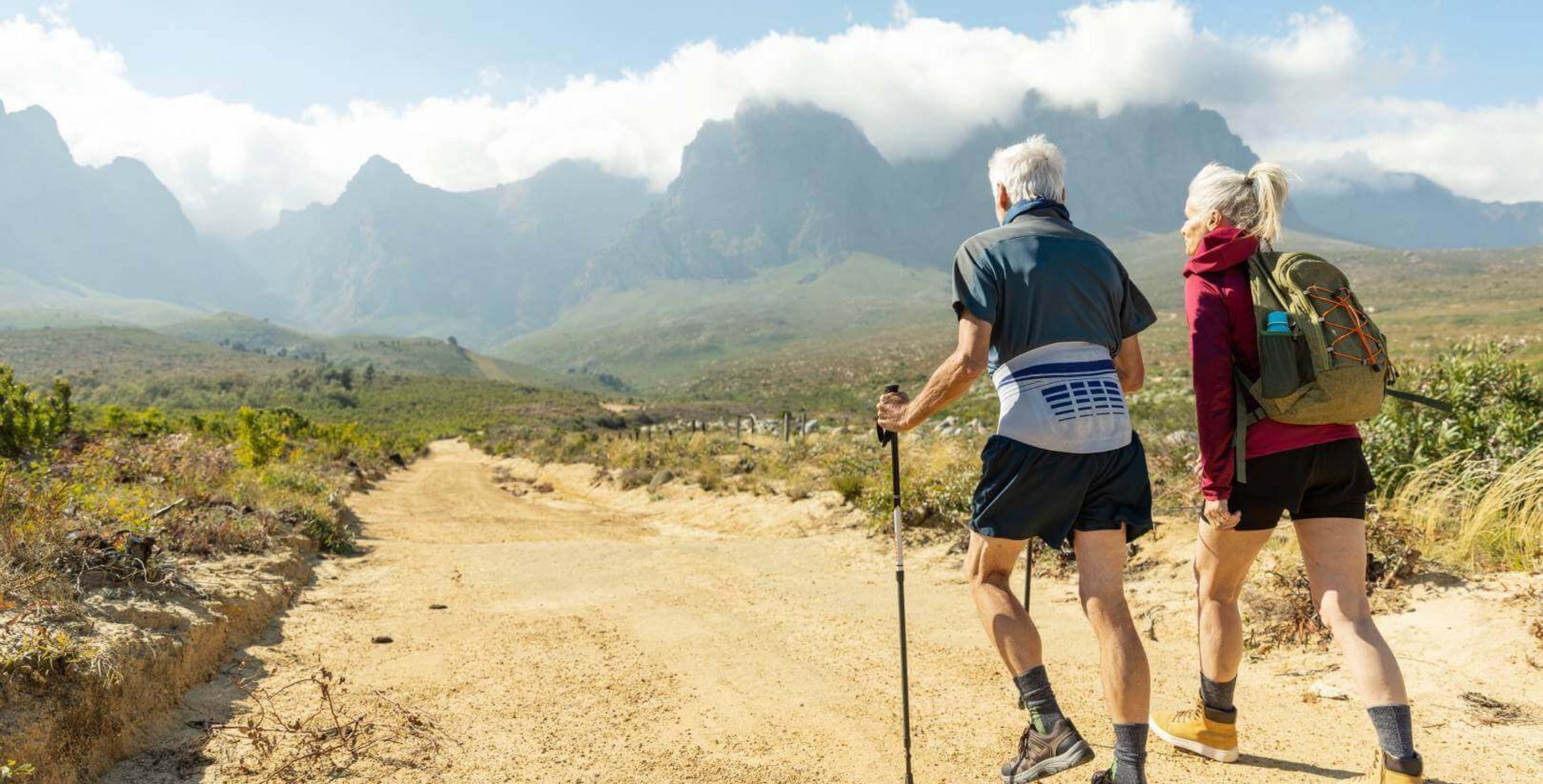 Abgebildet ist ein älteres Paar, das in einer sommerlichen Berglandschaft wandert. Zur Unterstützung nutzt der Mann Wanderstöcke und trägt eine Bauerfeind LumboLoc Rückenbandage.