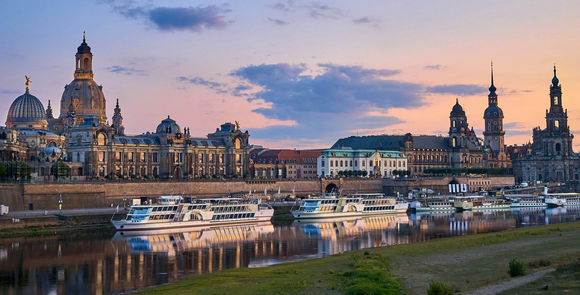 Abendliche Uferansicht der Dresdner Altstadt mit barocker Architektur, darunter die Frauenkirche und weitere historische Gebäude, spiegelnd im ruhigen Wasser der Elbe mit mehreren weissen Ausflugsschiffen am Ufer.
