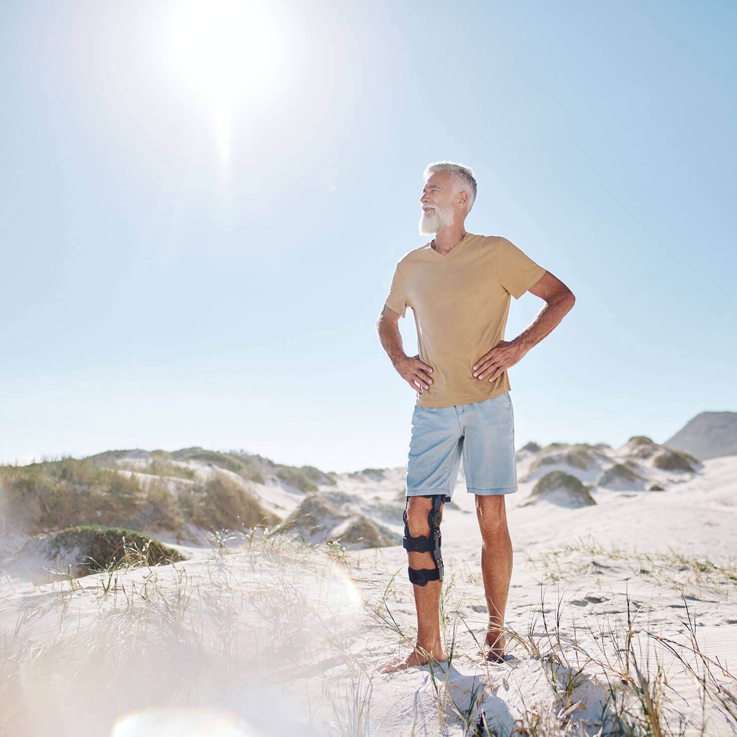 Ein Mann steht im Sand in einer Düne. Er trägt eine Orthese von Bauerfeind am rechten Knie, welche das Kniegelenk entlastet und stabilisiert. 