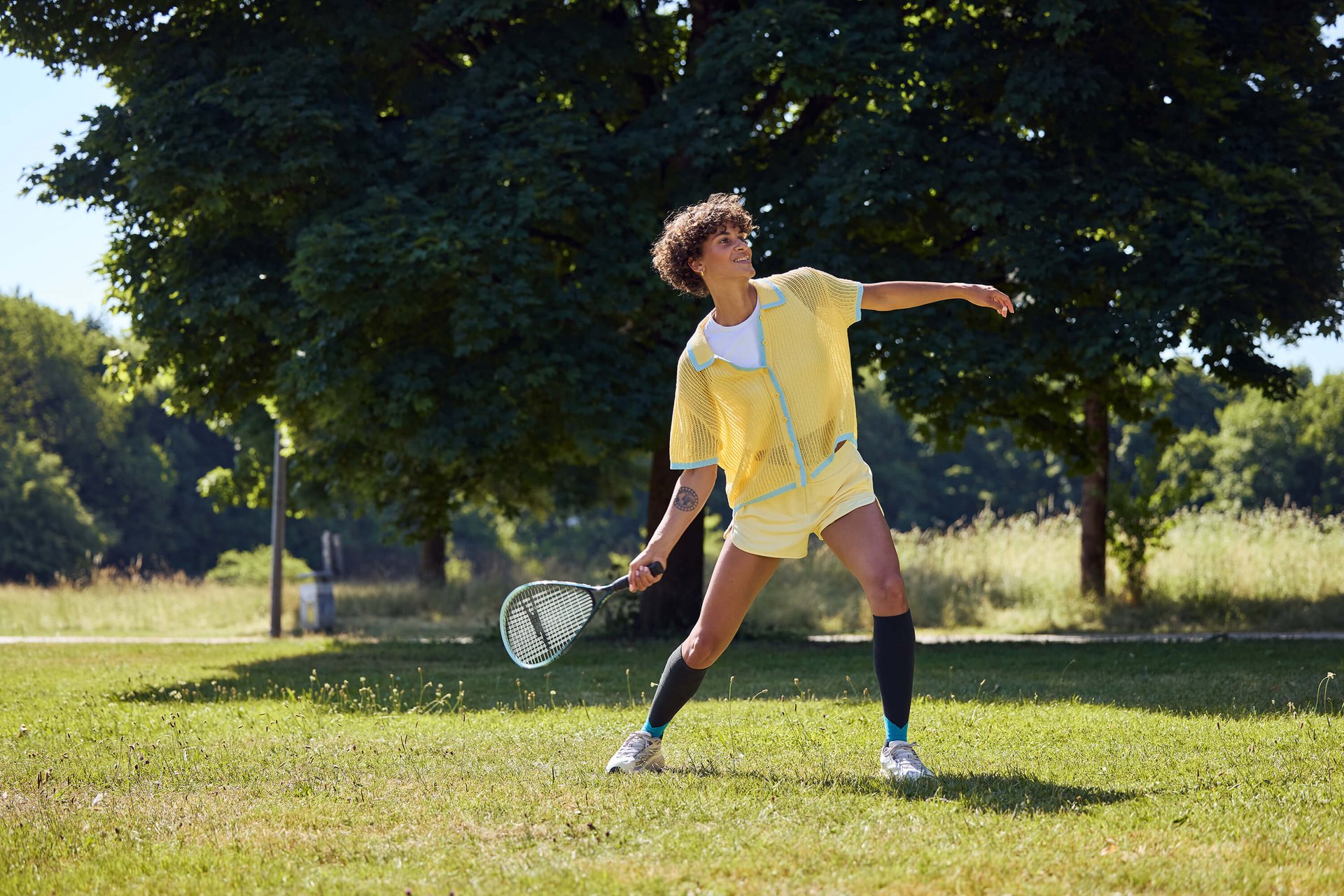 Person in gelbem Sportoutfit spielt mit einem Schläger auf einer sonnigen Wiese vor grossen Bäumen. Person in gelbem Sportoutfit spielt mit einem Schläger auf einer sonnigen Wiese vor grossen Bäumen.