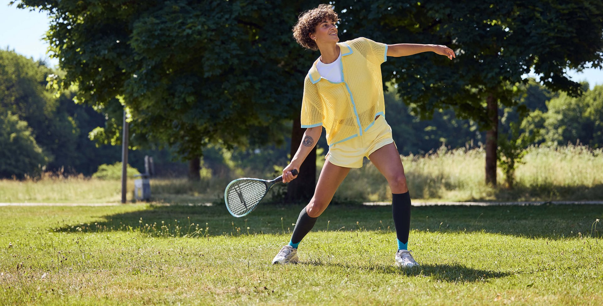 Person in gelbem Sportoutfit spielt mit einem Schläger auf einer sonnigen Wiese vor grossen Bäumen.