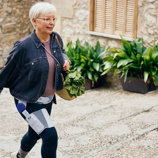 Eine ältere Frau mit kurzen weissen Haaren und Brille läuft auf einer gepflasterten Strasse entlang. Sie trägt eine Jeansjacke, ein rosafarbenes Oberteil und dunkle Leggings, an denen medizinische Geräte befestigt sind. In der Hand hält sie einen Korb mit frischem Grünzeug. Im Hintergrund sind eine Steinwand mit Fensterläden und grüne Pflanzen in Töpfen zu sehen.