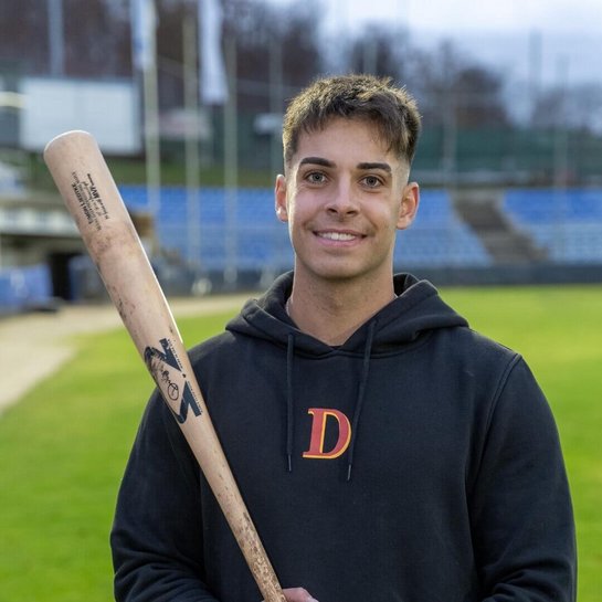 Simon Liedkte, Baseballspieler, steht lächelnd auf einem Baseballplatz. Er trägt eine schwarze Kapuzenjacke mit einem roten "D"-Logo und hält einen Baseballschläger locker über die Schulter. 