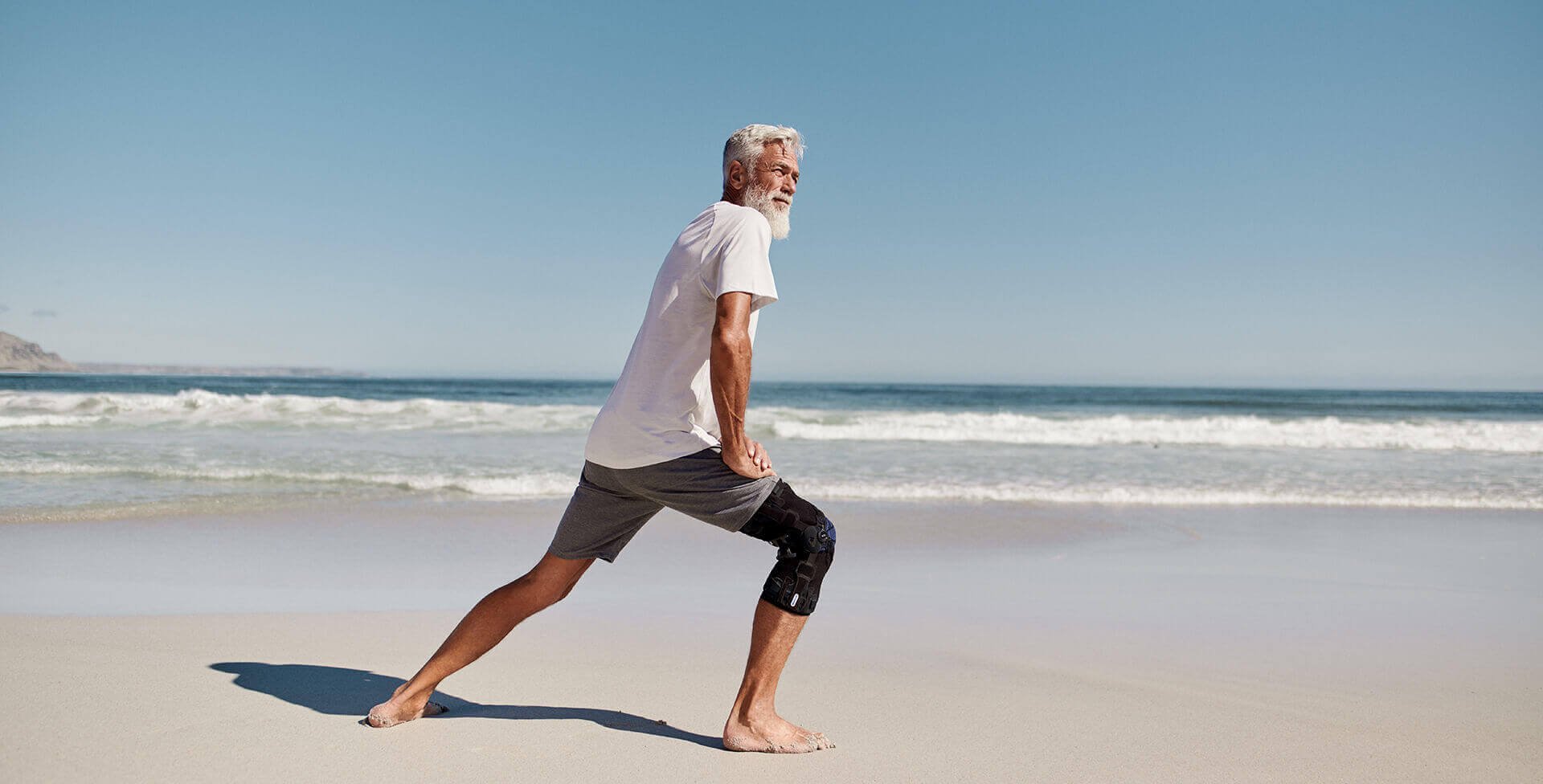 Ein Mann ist am Strand und macht mit den rechten Bein einen Ausfallschritt nach vorn und stützt dabei seine Hände auf den rechten Oberschenkel ab. Er trägt eine GenuTrain OA Kniebandage von Bauerfeind für eine gezielte Entlastung und Stabilisierung bei Kniearthrose.