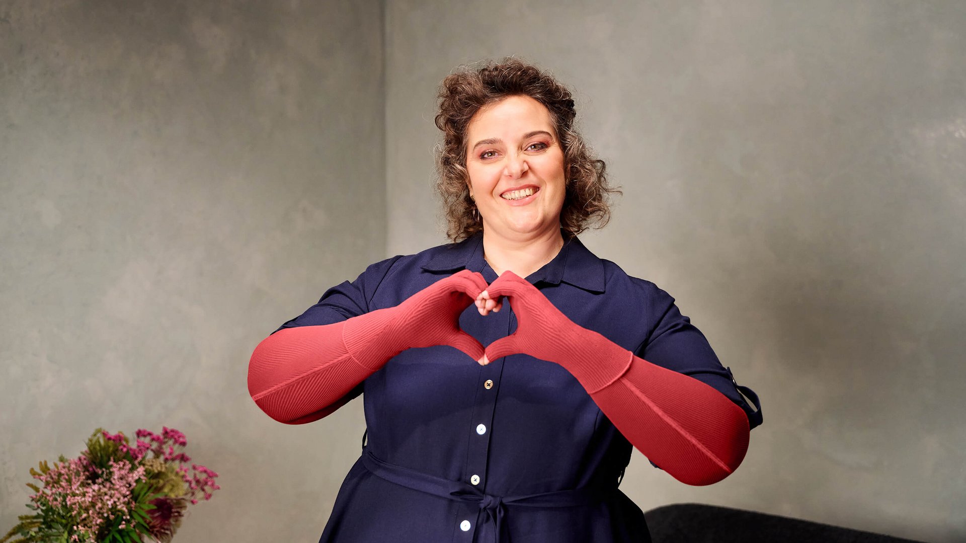 Eine lächelnde Frau steht vor einer hellgrauen Wand und formt mit ihren roten Kompressionshandschuhen ein Herz vor ihrer Brust. Links im Bild ist ein Blumenstrauss zu sehen.