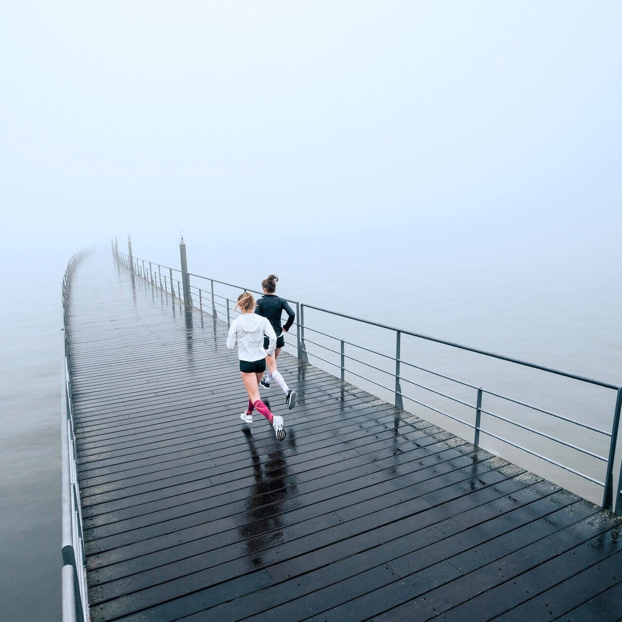 Zwei junge Frauen laufen über einen Steg am Ufer entlang. Sie tragen Run Performance Compression Socks von Bauerfeind, welche die Wadenmuskulatur durch gezielte Kompression unterstützen.