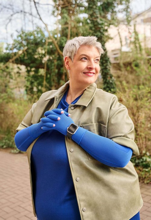 Ältere Frau mit kurzen grauen Haaren steht draussen auf einem gepflasterten Weg und lächelt zur Seite. Sie trägt ein blaues Oberteil mit dazu passenden blauen Handschuhen, eine olivgrüne Jacke sowie eine Smartwatch am linken Handgelenk. Im Hintergrund sind Bäume und Kletterpflanzen zu sehen.