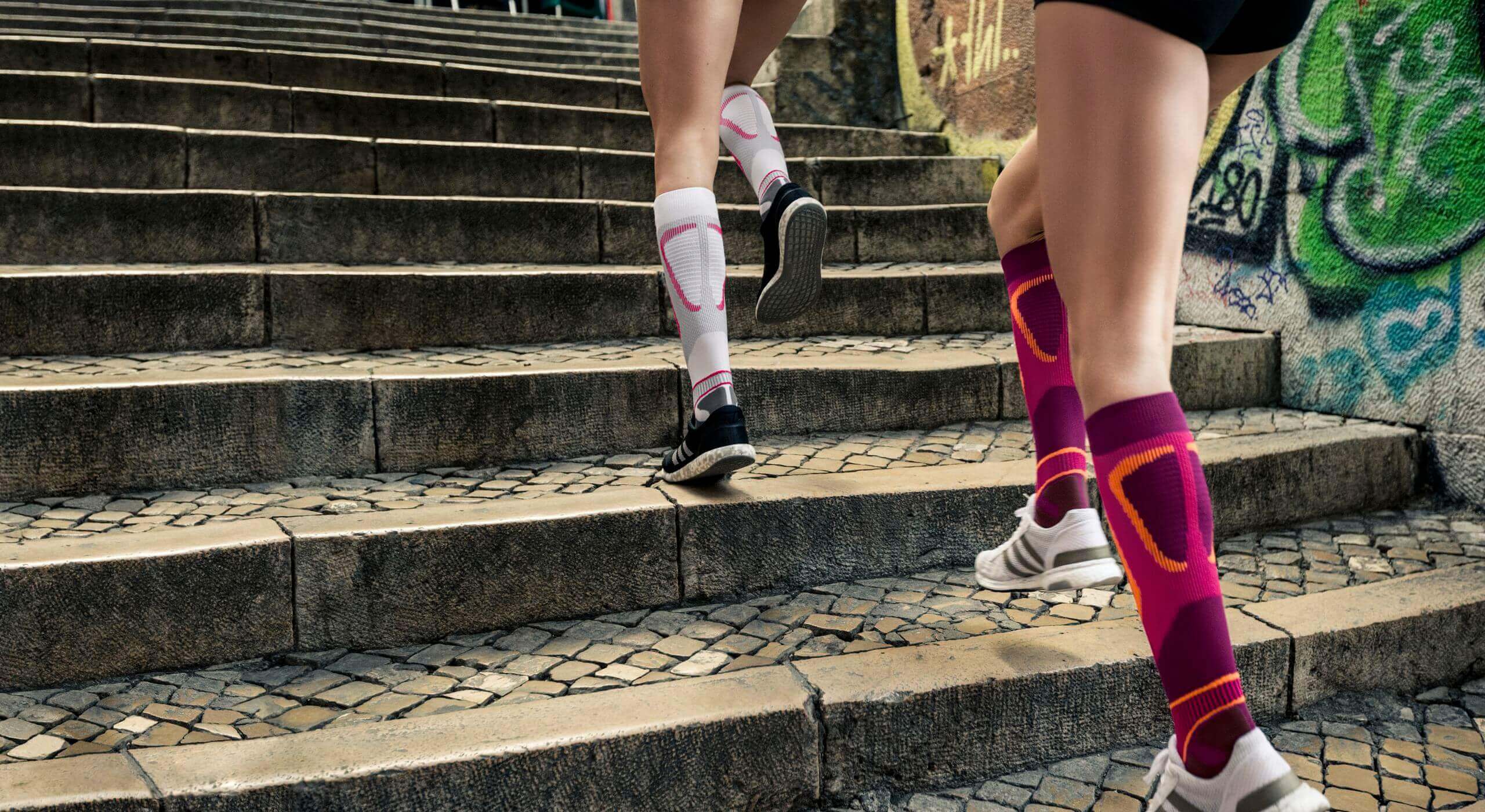 Bildauschnitt zweier Frauen, die eine Treppe hochrennen. Sie tragen beide Run Performance Compression Socks von Bauerfeind. welche die Wadenmuskulatur durch gezielte Kompression unterstützen.