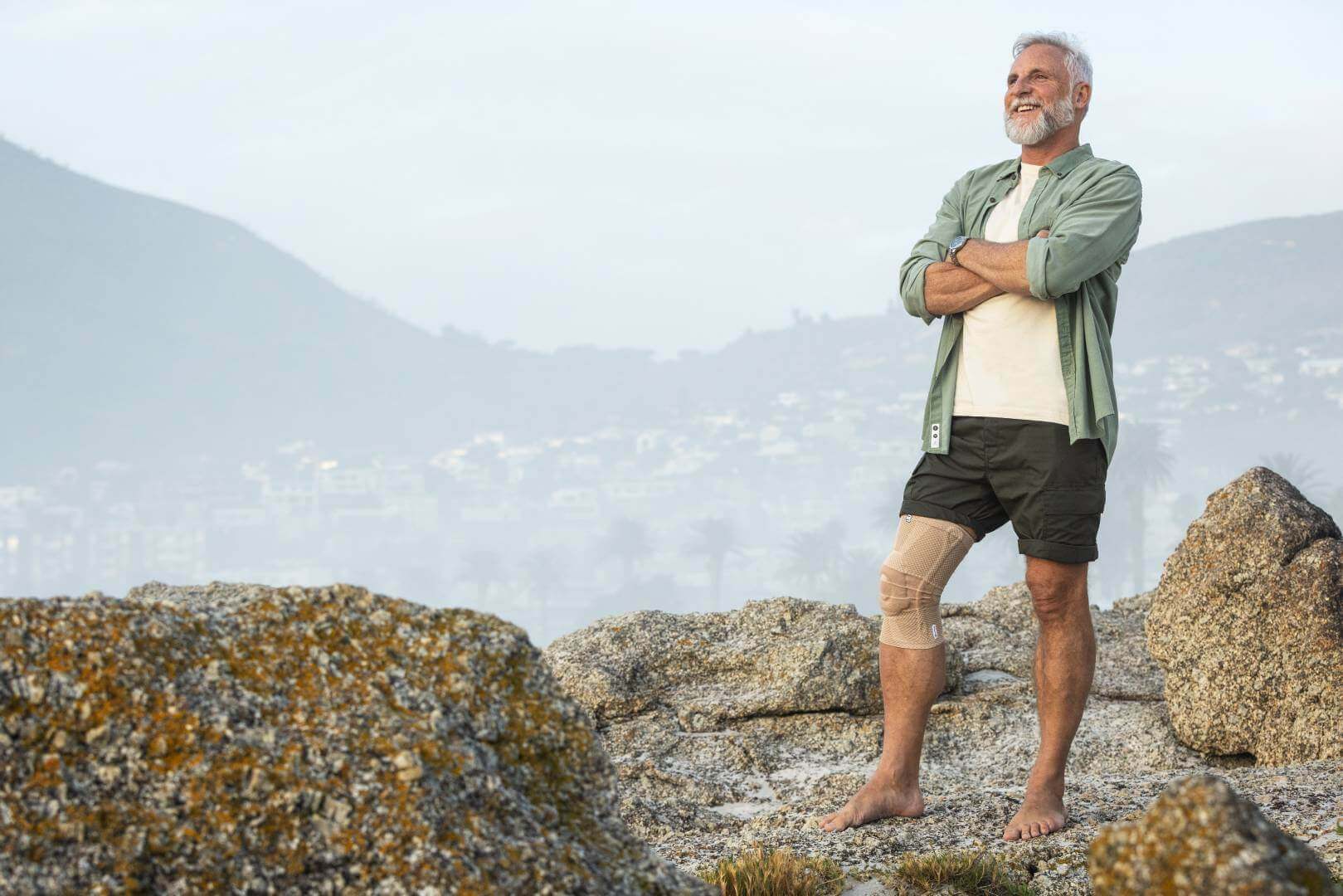 Das Bild zeigt einen älteren Mann, der an der Küste steht und den Ausblick genießt. Am Knie hat er eine GenuTrain Bandage zur Linderung von arthrosebedingten Schmerzen.